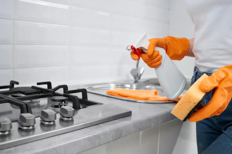 Gloved hands deep cleaning a kitchen countertop and stovetop with cleaning spray